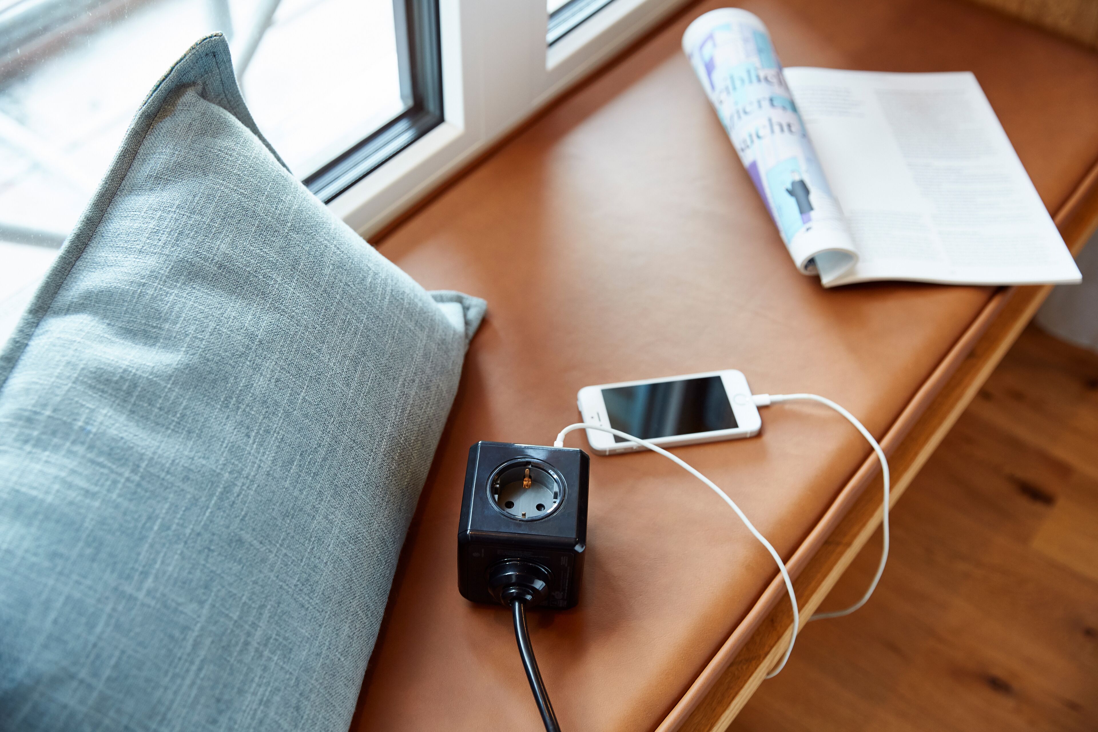 Eine braune Lederbank mit einem Smartphone, einem Ladekabel, einer aufgerollten Zeitschrift und einem grauen Kissen.