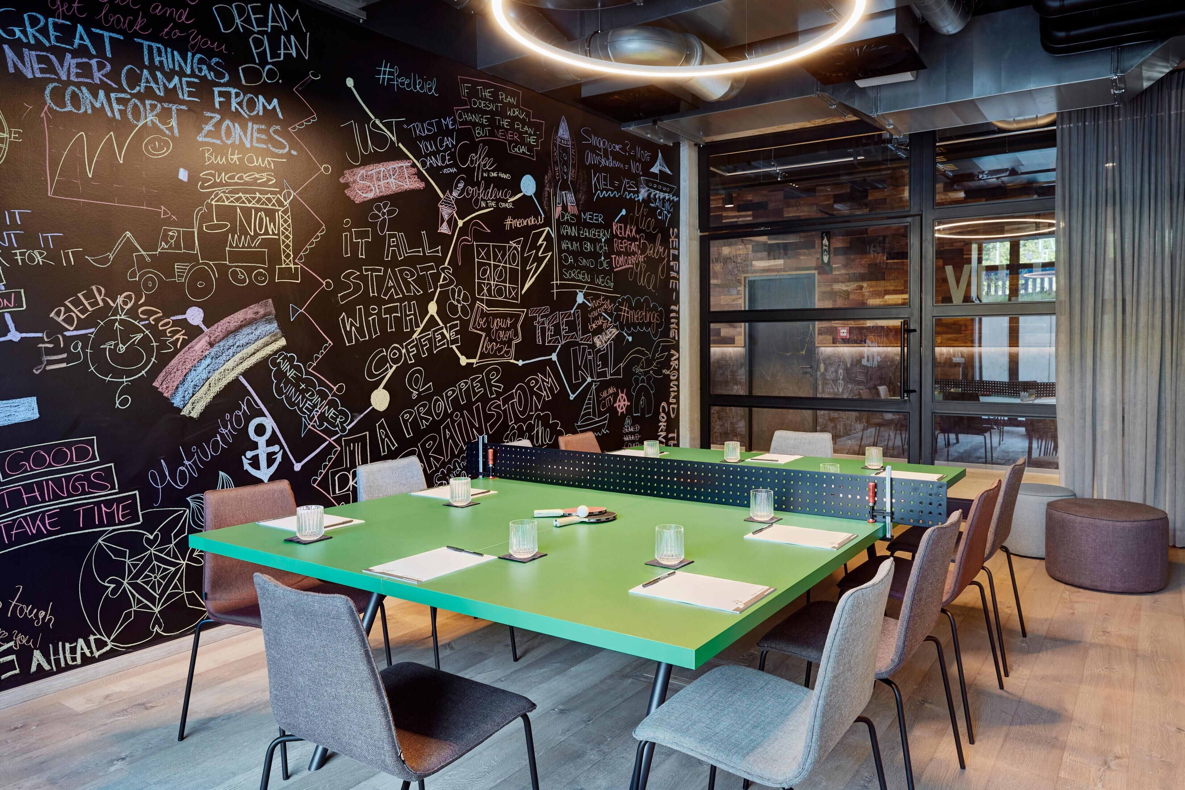 Ein moderner Konferenzraum mit einem grünen Tisch, weißen Stühlen und einer Tafel mit motivierenden Sprüchen und Kritzeleien.