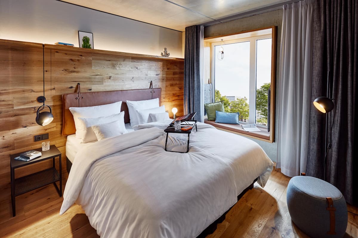 Une chambre d’hôtel moderne avec une tête de lit en bois, un grand lit avec du linge de lit blanc, une table de chevet avec une lampe et une vue sur l’extérieur par une fenêtre.