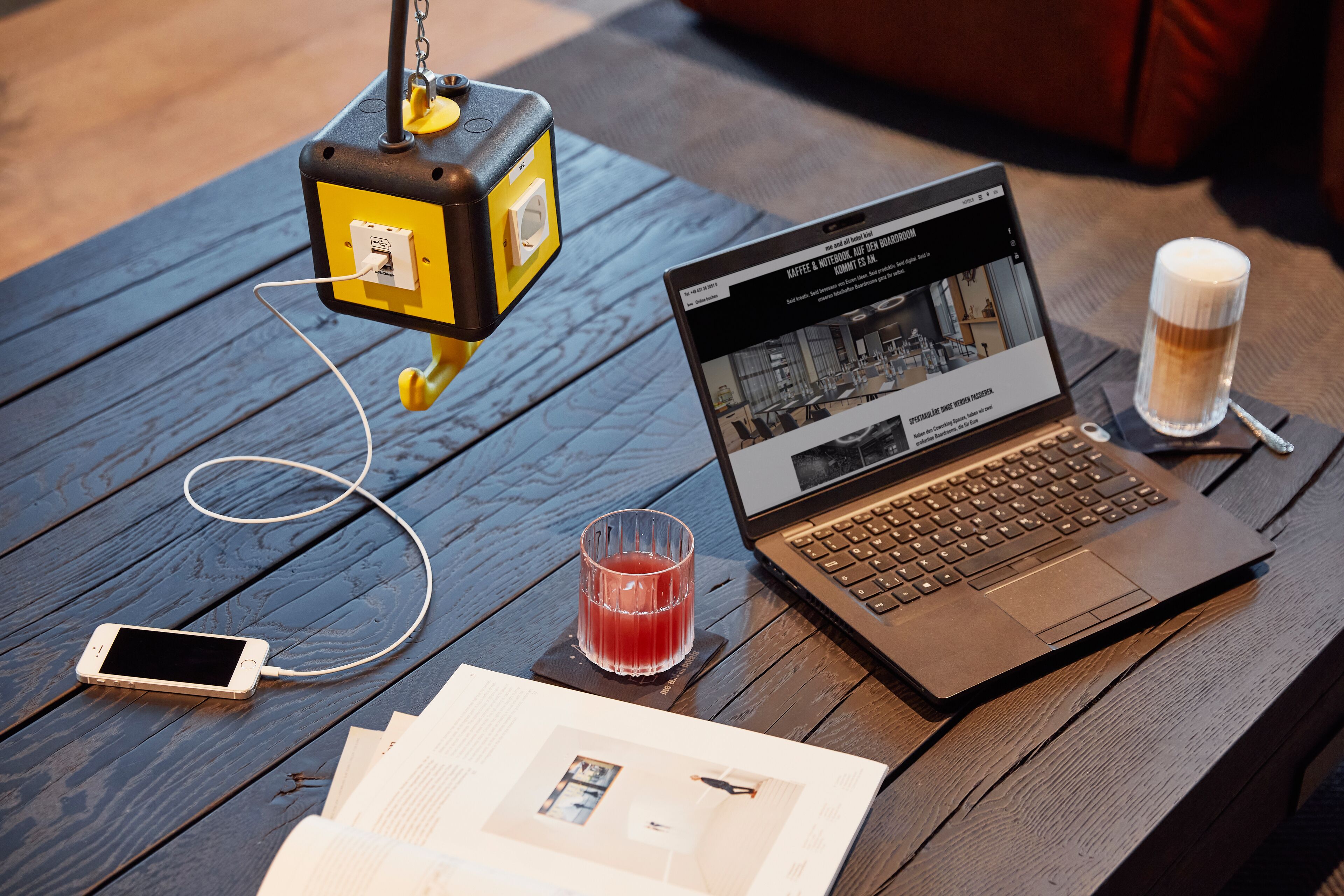 A laptop on a wooden table with a smartphone, a glass of red drink, and a power strip.