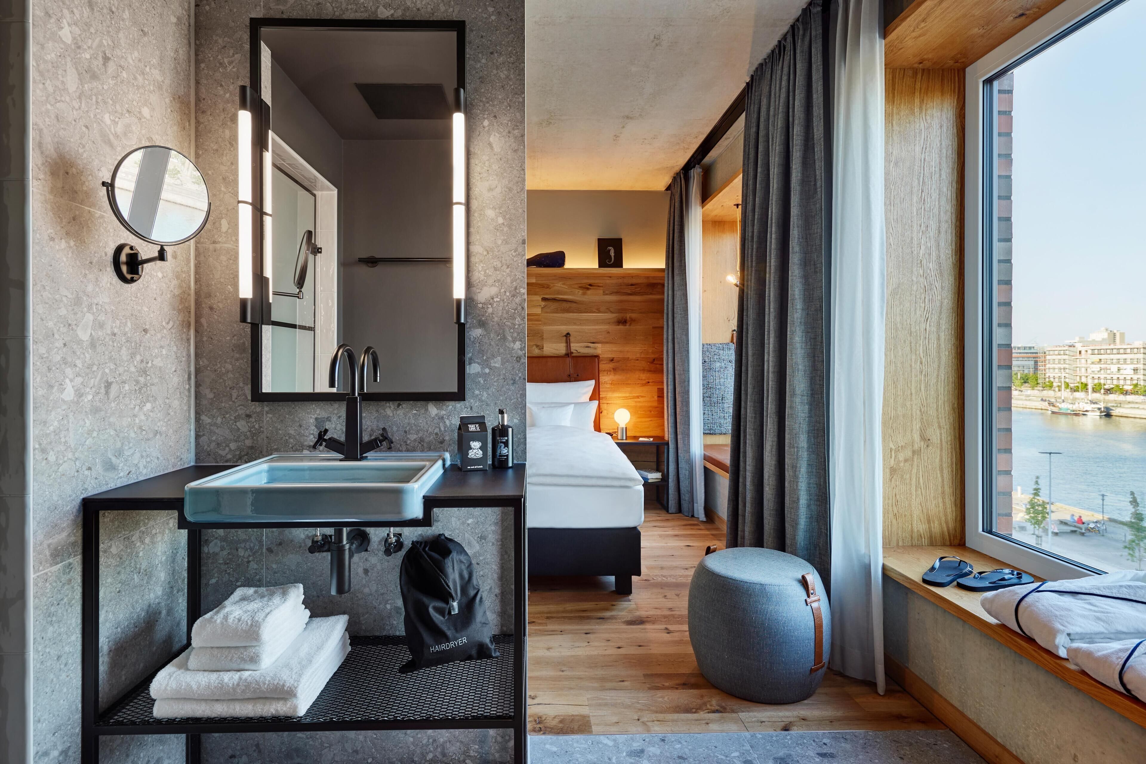 Una habitación de hotel moderna con un ventanal grande, una cama, un baño con lavamanos y espejo, y un pequeño espacio de descanso.