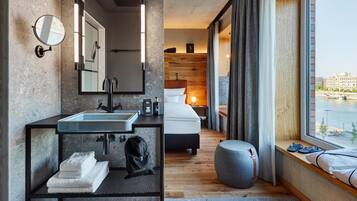 Una habitación de hotel moderna con un ventanal grande, una cama, un baño con lavamanos y espejo, y un pequeño espacio de descanso.