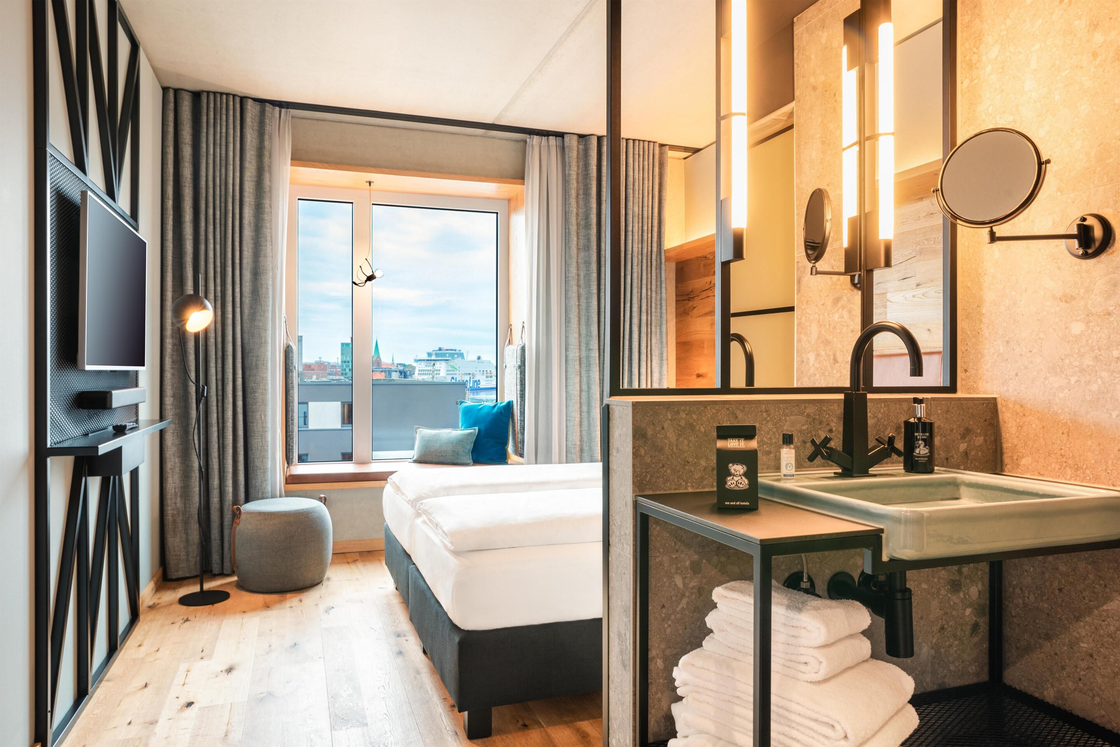 Une chambre d’hôtel moderne équipée d’un lit, d’une grande fenêtre, d’une télévision et d’une salle de bain avec un lavabo et un miroir, offrant une vue sur la ville.