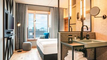 Une chambre d’hôtel moderne équipée d’un lit, d’une grande fenêtre, d’une télévision et d’une salle de bain avec un lavabo et un miroir, offrant une vue sur la ville.