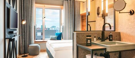 Una habitación de hotel moderna con cama, un ventanal amplio, televisión, baño con lavamanos y espejo, y vistas a la ciudad.