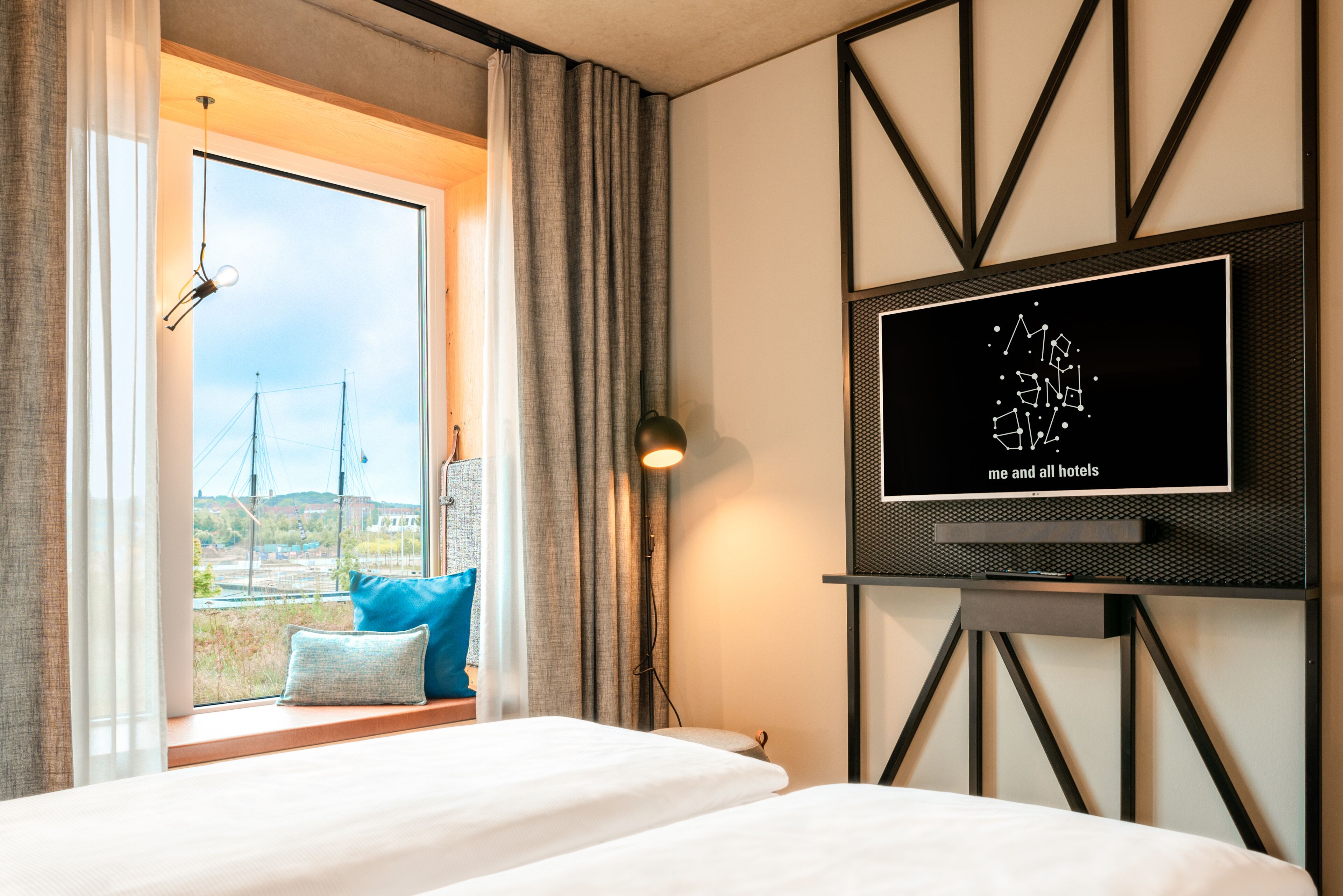 Une chambre d’hôtel avec une grande fenêtre, un lit avec une literie bleue et blanche, un téléviseur fixé au mur et une vue sur un pont et des immeubles à l’extérieur.