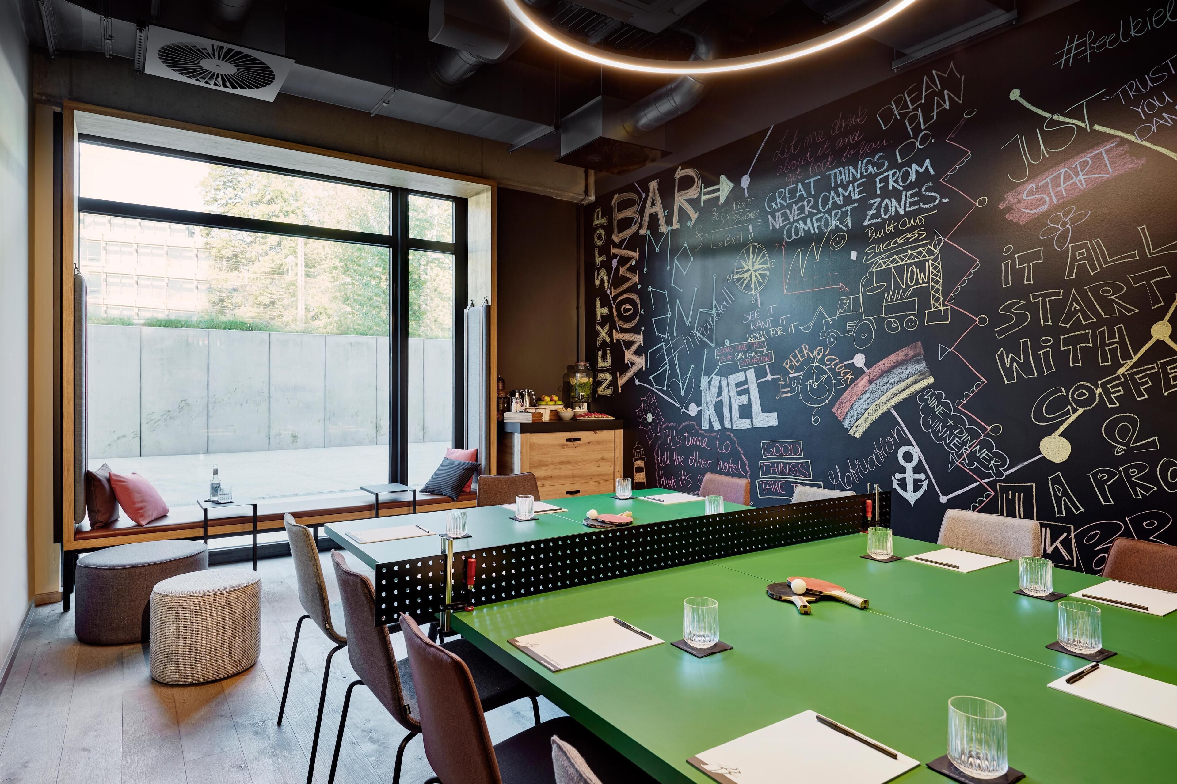 Una sala de reuniones moderna con una mesa verde, sillas blancas, un ventanal grande y un mural motivacional.