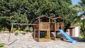 Une structure de jeu en bois, dotée d’un toboggan et de balançoires, installée sur une terrasse en pierre avec des arbres en arrière-plan.