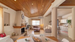 Un vaste espace de vie doté d’un plafond en bois, d’une cheminée, d’une table à manger avec des chaises, et d’une cuisine située à l’arrière-plan.