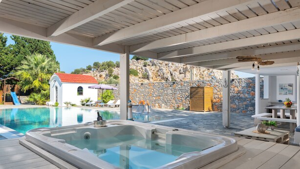 Un espace piscine comprenant un jacuzzi, une terrasse en bois, un mur de pierres et un petit bâtiment blanc avec un toit rouge.