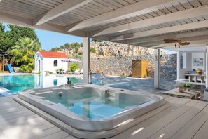 A pool area with a hot tub, wooden deck, stone wall, and a small white building with a red roof. - Madia Villa in Kolympia (Kolympia)