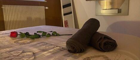Un lit bien fait, avec des serviettes pliées en rouleaux, une rose rouge et un mur décoratif orné d’un panneau indiquant le mot « amour ».