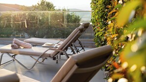 Un área de descanso al aire libre iluminada por el sol, con dos sillones reclinables, una toalla enrollada y una barandilla de vidrio con vistas a un jardín y un paisaje lejano.