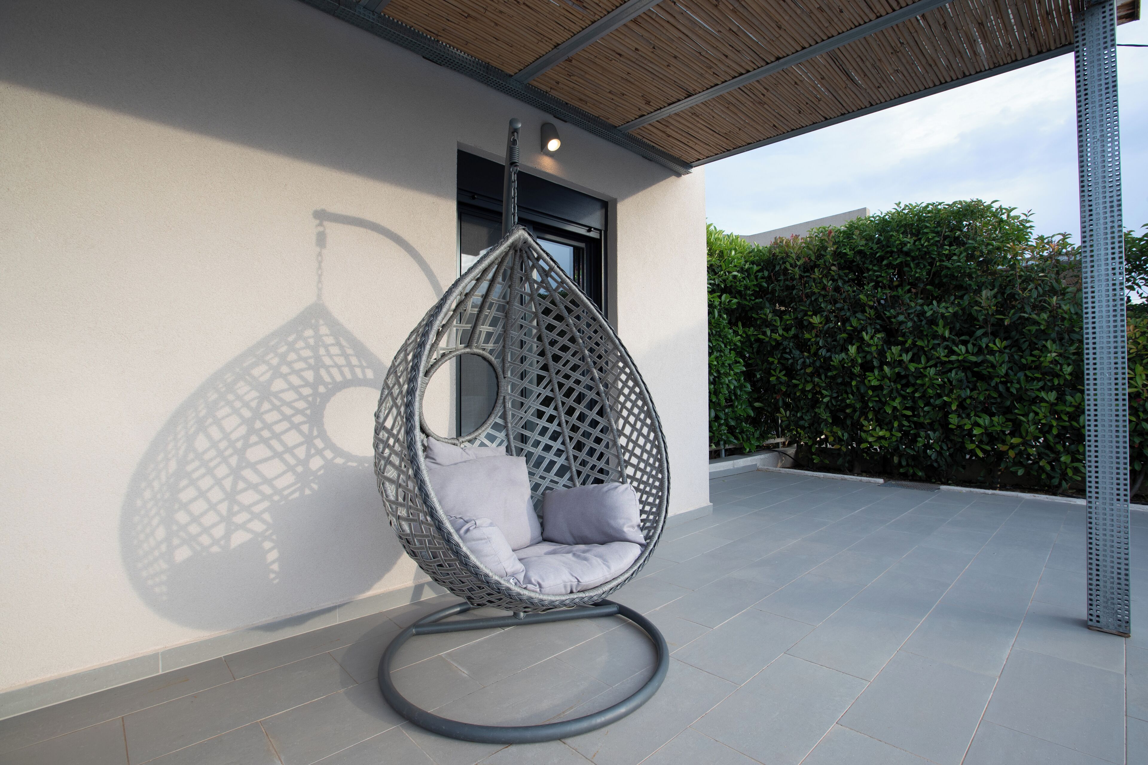 Un fauteuil suspendu doté d’une structure circulaire et de coussins, placé sur une terrasse carrelée avec une rambarde en métal et un mur percé d’une fenêtre.