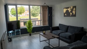 A modern living room with a large window, a flat-screen TV, a dark grey sofa, a small coffee table, and a potted plant.
