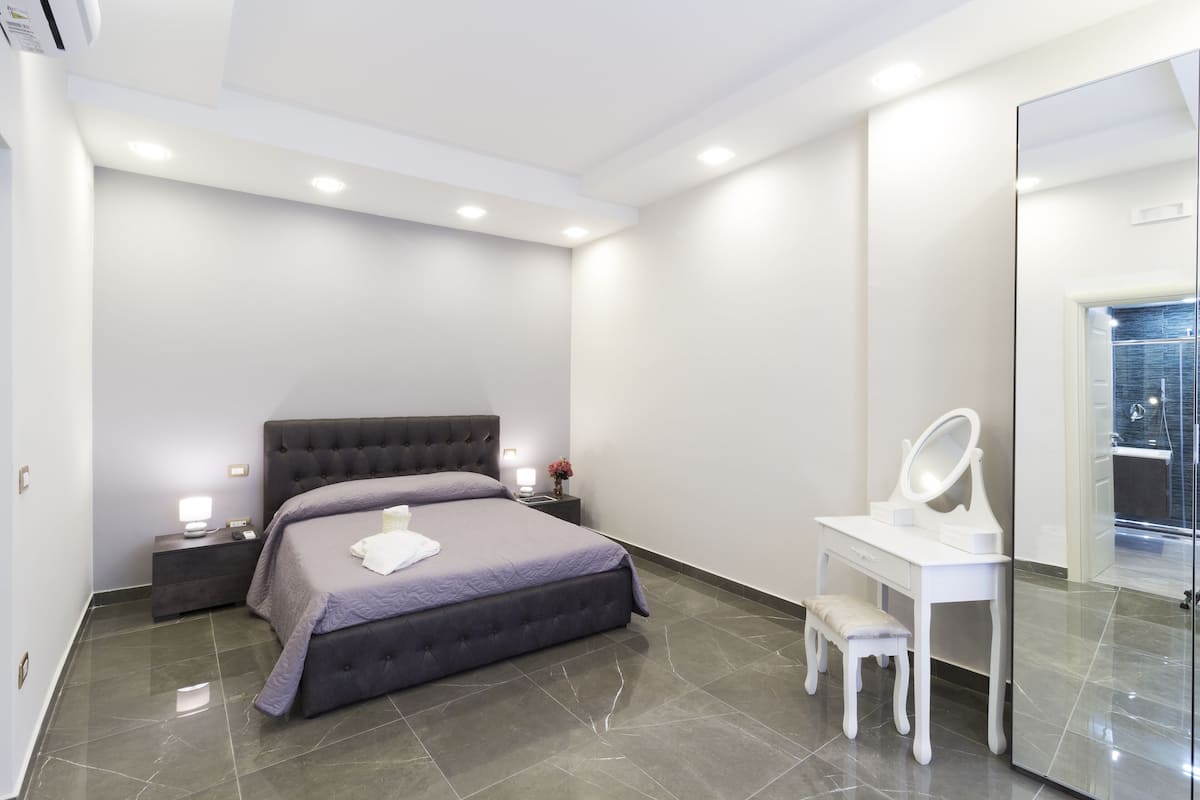 A modern bedroom with a large bed, bedside tables, a vanity with a mirror, and a shower area.