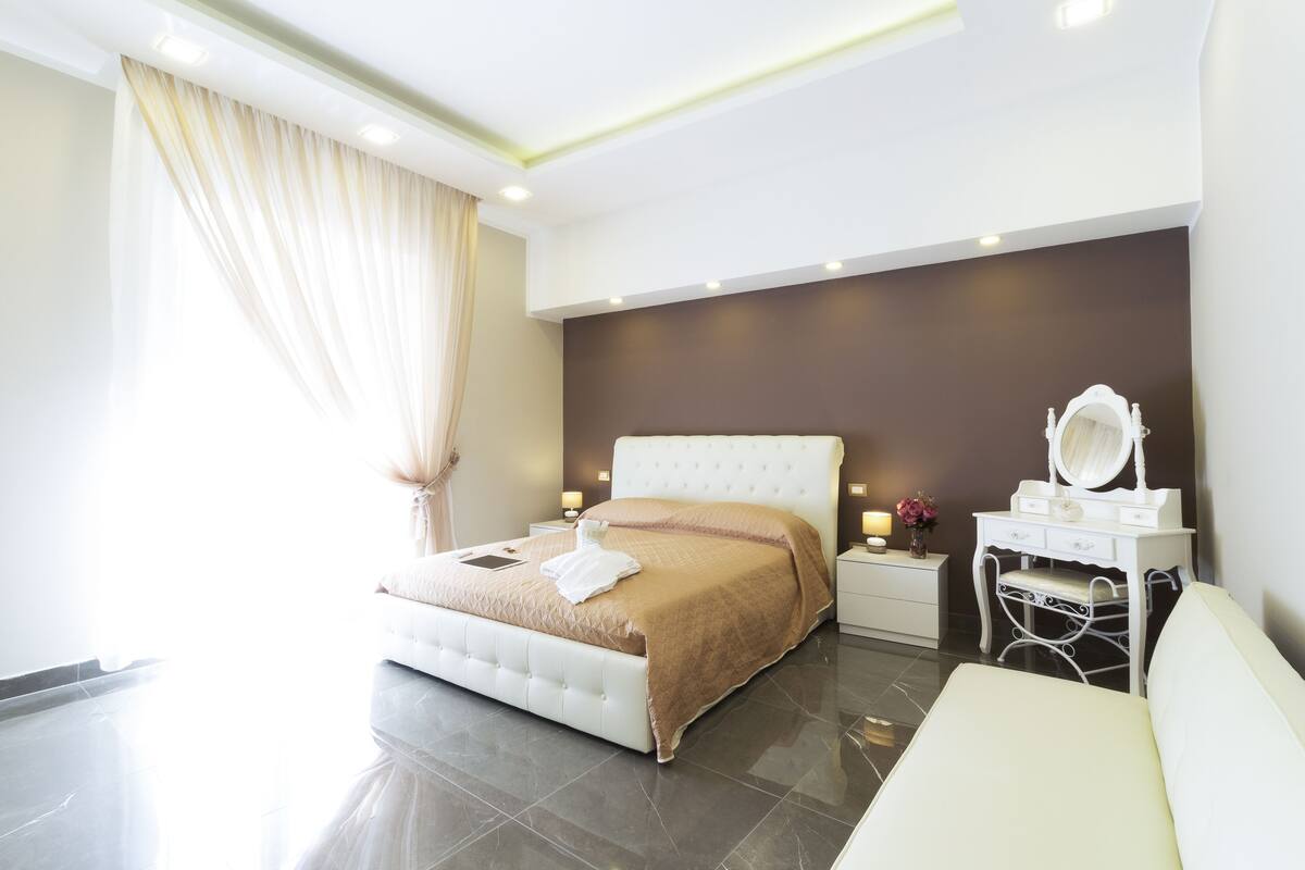A modern bedroom with a large bed, bedside tables, a vanity with a mirror, and a white sofa.