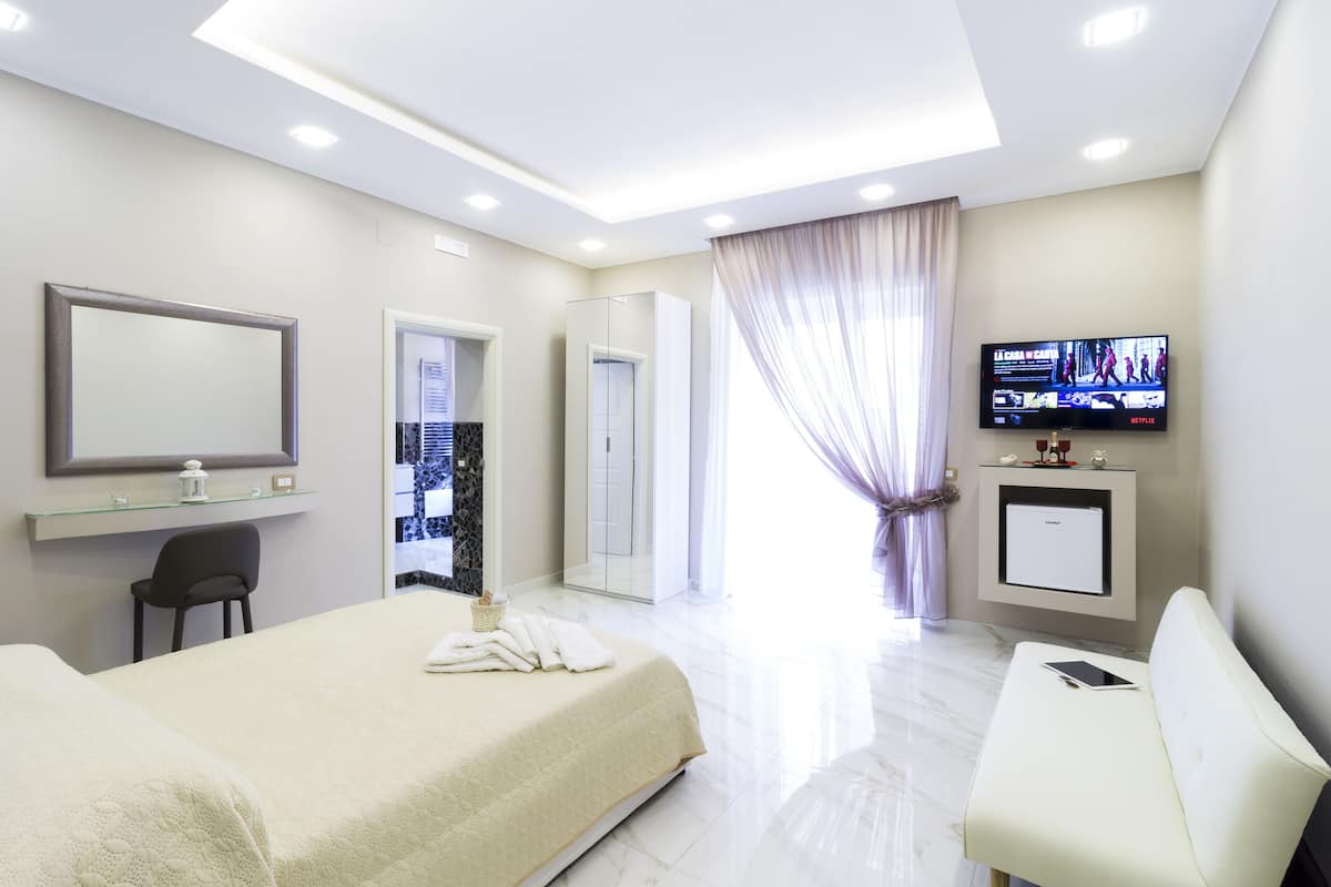 A modern hotel room with a large bed, a desk, a chair, a TV, and a bathroom with a shower and toilet.
