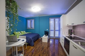 A compact room with a bed, a desk with a laptop, a small table, a kitchen area, and a window with blinds. - Aurum ApartHotels Pianell (Milan)