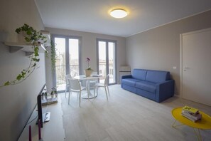 A modern living room with a blue sofa, a white dining table with chairs, a yellow side table, and a balcony with a view. - Aurum ApartHotels Pianell (Milan)