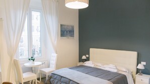 A bedroom with a bed, bedside tables, a chair, a small table, and a window with curtains. - Vatican Garden Rooms (Rome)