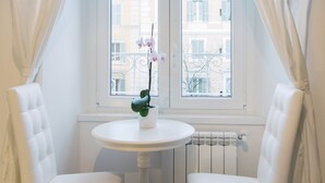 A room with a white round table, two white chairs, a potted orchid, and a view of buildings through a window with white curtains. - Vatican Garden Rooms (Rome)