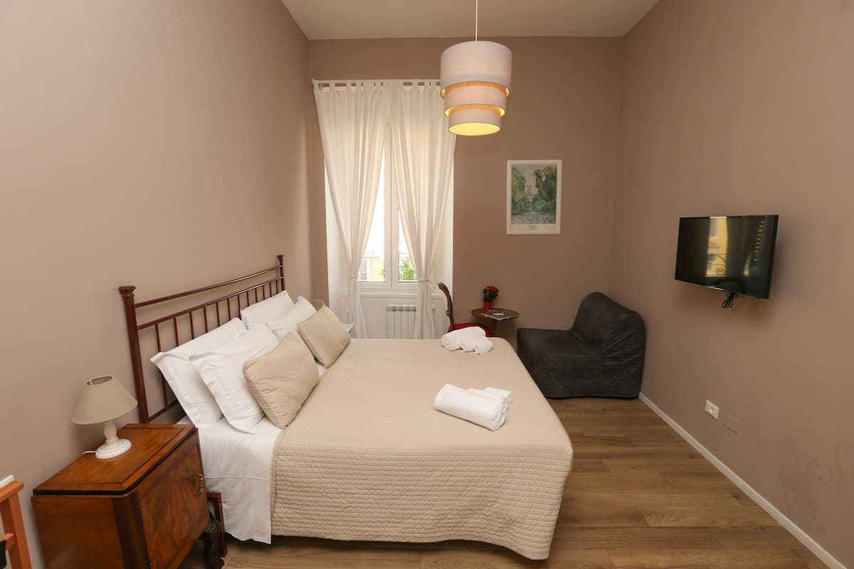 Une chambre à coucher avec un lit, une table de chevet, une lampe, des rideaux, une chaise, un téléviseur et un tableau accroché au mur.