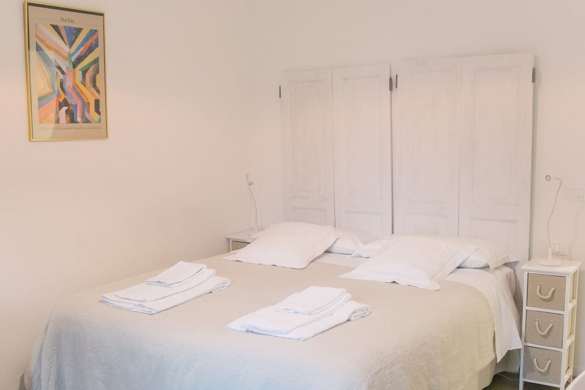 Un lit bien fait, avec du linge de lit blanc, une table de chevet avec une lampe et une œuvre d’art accrochée au mur.