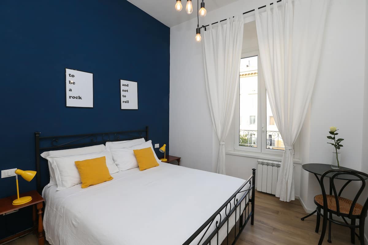 Une chambre à coucher comprenant un lit, une table de chevet, une lampe, une chaise, une table et une fenêtre avec des rideaux.