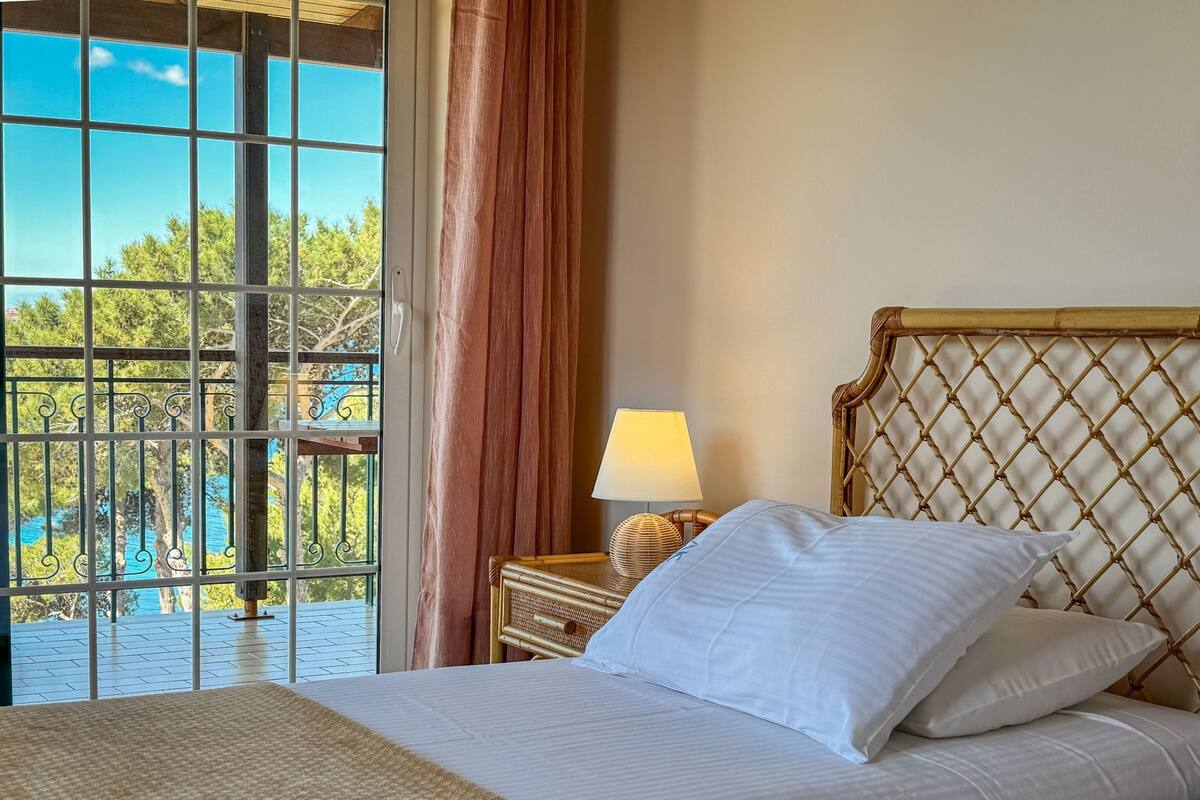 A hotel room with a bed, a nightstand, a lamp, and a view of a pool and greenery.