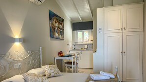 A bedroom with a bed, pillows, a bedside table, a small desk, and a kitchenette in the background. - Anthoula's House (Kefalonia)