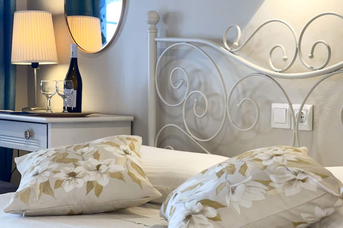 A neatly made bed with floral pillows, a bedside table with a wine bottle and glasses, and a wall-mounted light fixture.