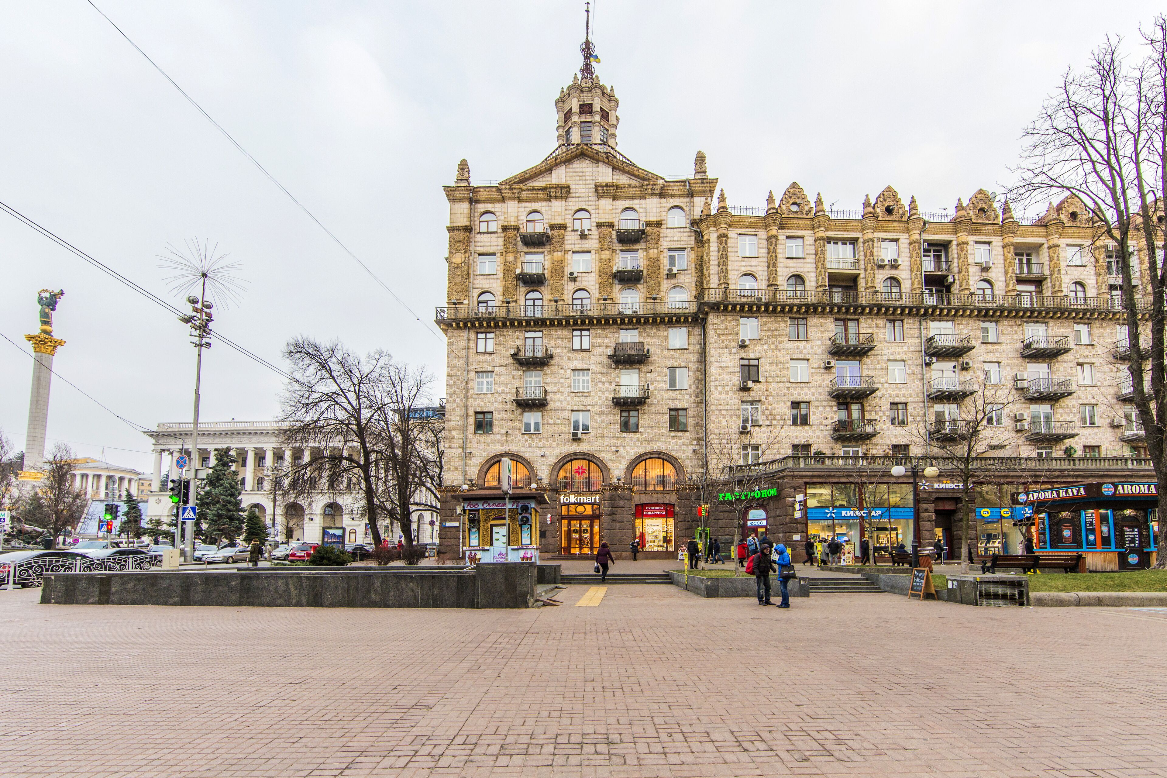 Un grand bâtiment historique, doté de plusieurs étages et de balcons, entouré d’une place pavée où se trouvent des bancs et quelques personnes.