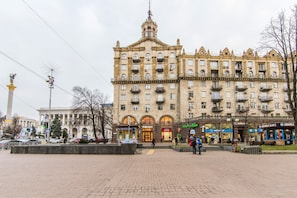 Un grand bâtiment historique, doté de plusieurs étages et de balcons, entouré d’une place pavée où se trouvent des bancs et quelques personnes.