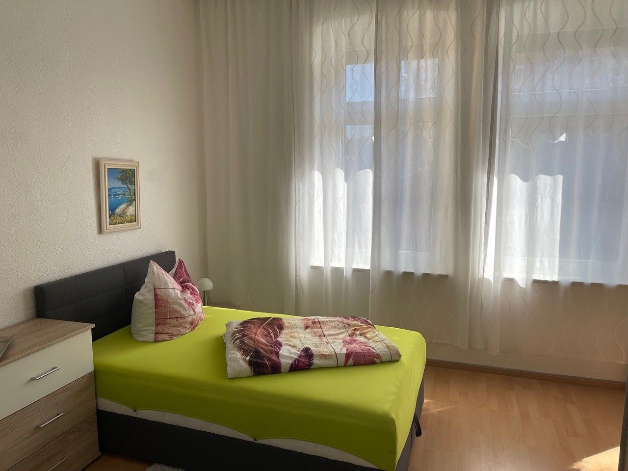 Ein Zimmer mit einem Bett, einem Nachttisch, einer Lampe, einem Bild an der Wand und einem Fenster mit durchscheinenden Vorhängen.