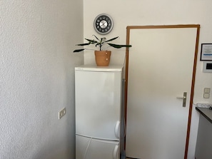 A compact room with a white refrigerator, a potted plant on top, a clock on the wall, and a door with a wooden frame. - Holiday apartment - private room at the Ginkgo tree (Erfurt)