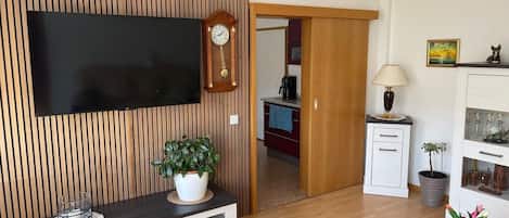 Una sala moderna con televisión de pantalla plana, pared de paneles de madera, un mueble pequeño, una lámpara y una planta en maceta.