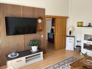 A modern living room with a flat-screen TV, wooden panel wall, a small cabinet, a lamp, and a potted plant.