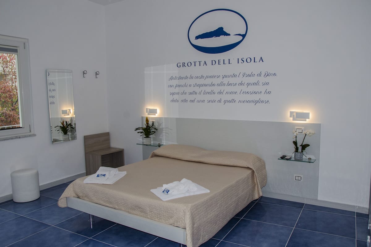 A hotel room with a bed, a mirror, a bedside table with a plant, and a wall-mounted sign with text.