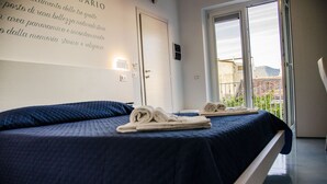 A hotel room with a bed, towels, a door, and a view of a building through a window. - Praja Vecchia (Praia a Mare)