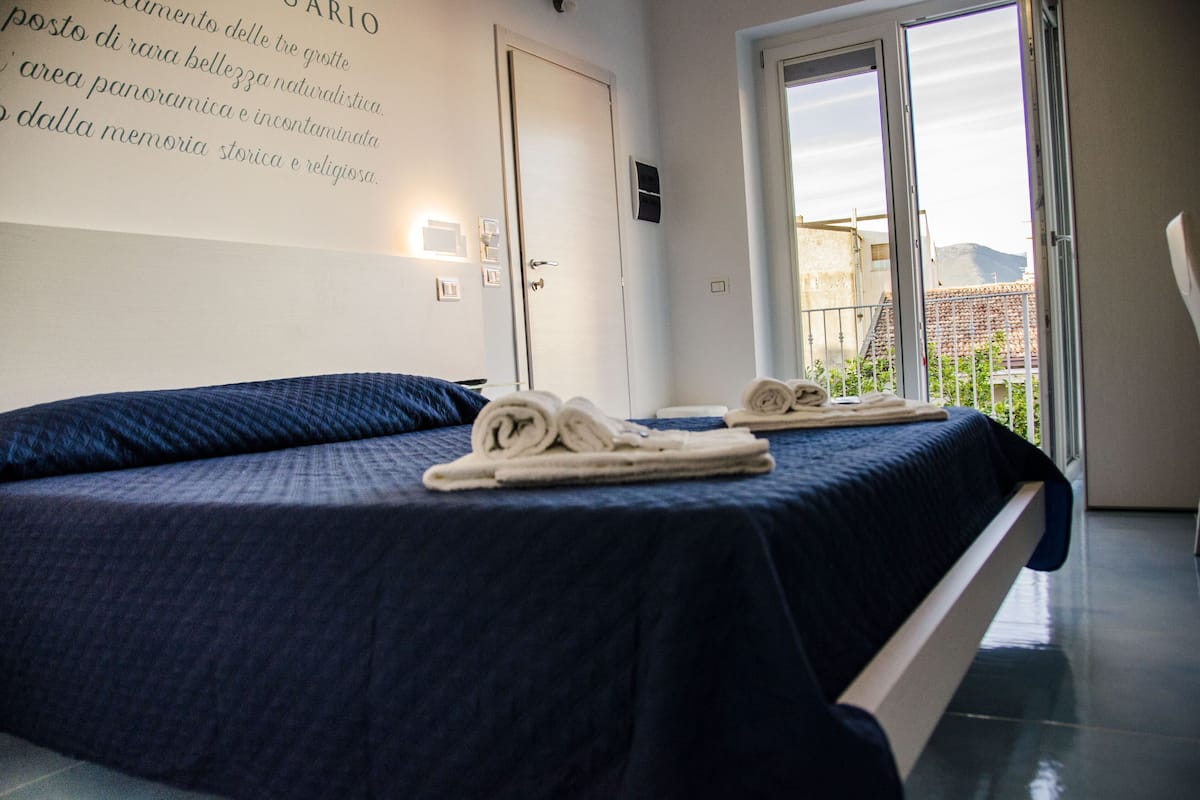 A hotel room with a bed, towels, a door, and a view of a building through a window.