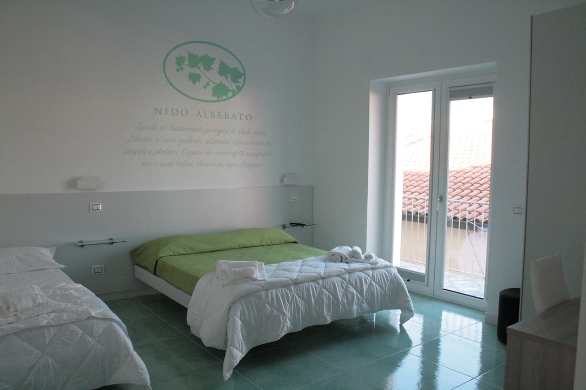 A hotel room with a bed, a green pillow, and a view of a red-roofed building through the door.