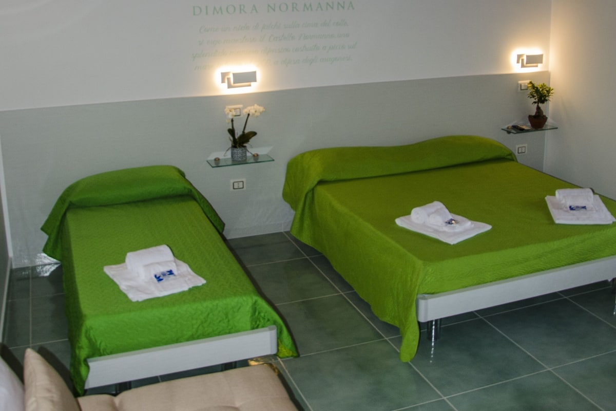 A room with two beds, each with a green bedspread and white towels. There is a small table with a vase of flowers and a wall-mounted light. The room has a wooden ceiling and tiled floor.
