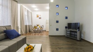 Ein Wohnzimmer mit einer Couch, einem Couchtisch mit Früchten, einem Fernseher und einem Essbereich im Hintergrund.
