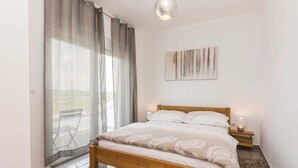 Une chambre à coucher avec un lit en bois, une table de chevet, une lampe fixée au mur et une fenêtre ornée de rideaux légers.