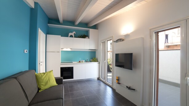 Un soggiorno moderno con un divano grigio, una TV a schermo piatto fissata al muro e un angolo cottura con mobili bianchi e una parete di accento blu.