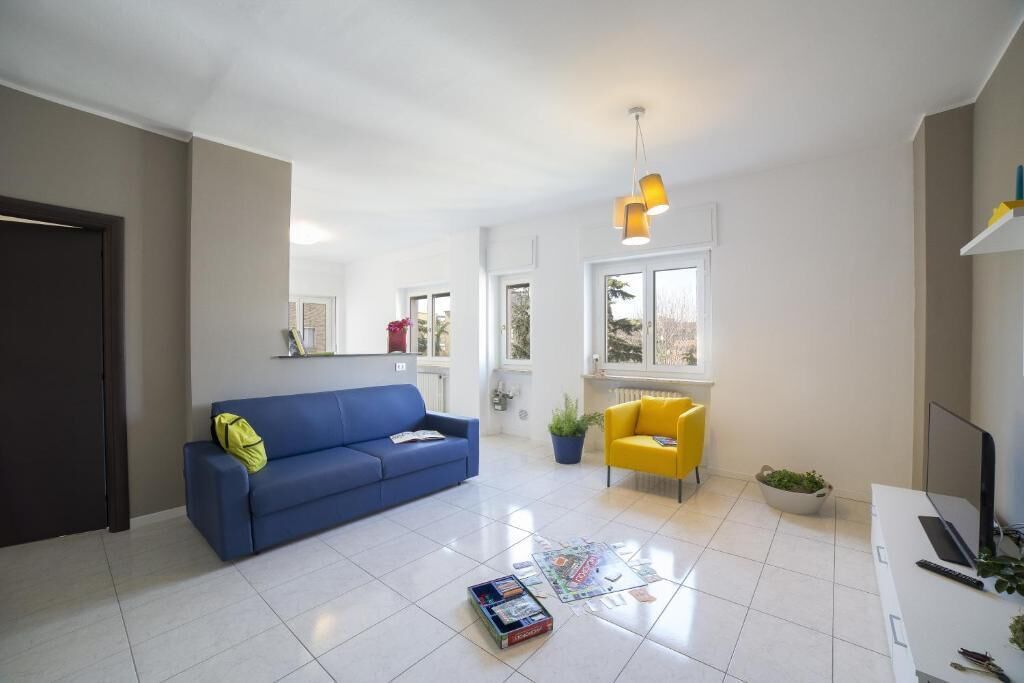 Un salon avec un canapé bleu, un fauteuil jaune, un bureau blanc avec un ordinateur portable et un luminaire suspendu.