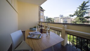 A balcony with a wooden table, a white chair, and a view of buildings and trees. - Aurum ApartHotels Gambara (Milan)