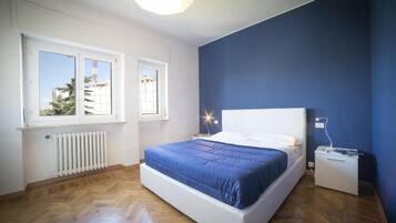 Un dormitorio con cama, dos ventanas, un radiador y una mesita de noche.