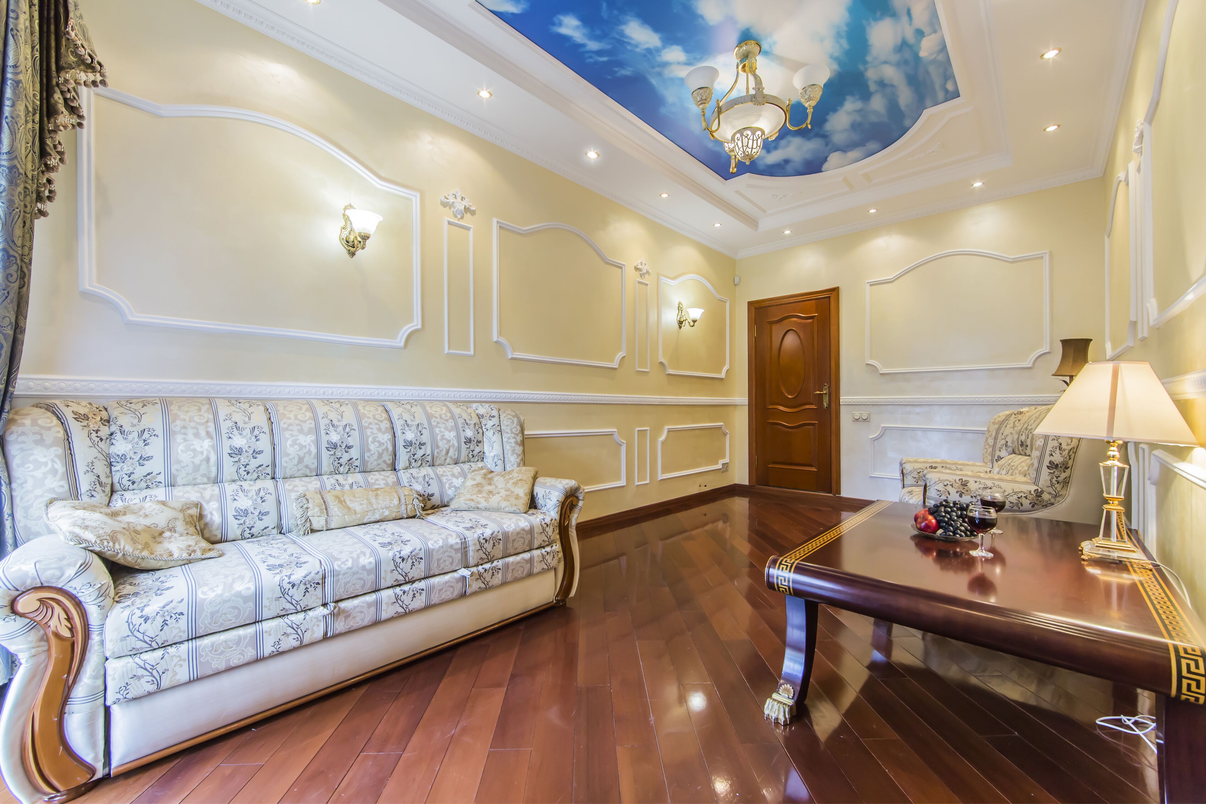 Una habitación amplia con suelo de madera, un sofá, una mesa con lámpara y un mural en el techo.